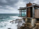 A modern luxury villa built into a rocky coastline, featuring saline-resistant metal frames and expansive glass overlooking a rough ocean.